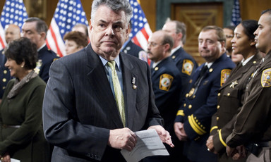 Peter King, a member of Congress for New York, stands beside US flags and soldiers in uniform.