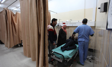 A man in medical scrubs and a man and woman in normal dress stand over a man lying on his side on a hospital bed