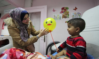 A young woman greets a child holding a balloon as he sits up on his hospital bed. 