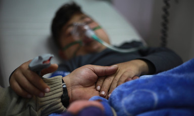 Young patient on a hospital bed, with a mask on his face and his hand being held by another person.