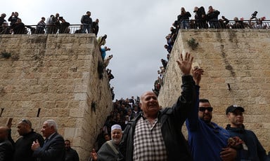 A man raising his hand surrounded by crowds with Bab al-Rahma gate in the background. 