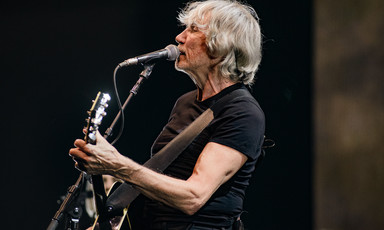 Roger Waters singing through a microphone with guitar. 