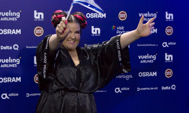 Woman dressed in black waves a blue and white flag in front of a backdrop featuring corporate logos.