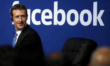 Man wearing ear piece stands in front of Facebook sign