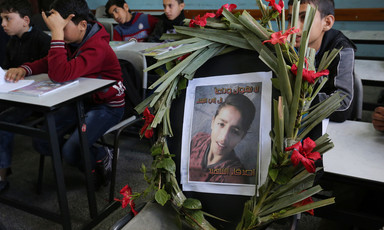 A wreath containing a large picture of a boy inside it is propped up on a chair; children sit near the wreath in a classroom.