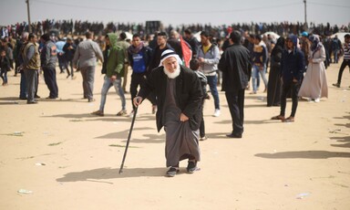 Man wearing white headdress walks with aid of stick surrounding by various people. 