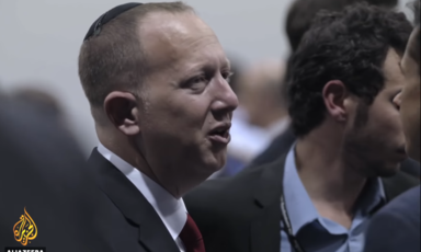 A man in a suit and red tie, wearing a kippah, a Jewish skull cap, talks to two other men.