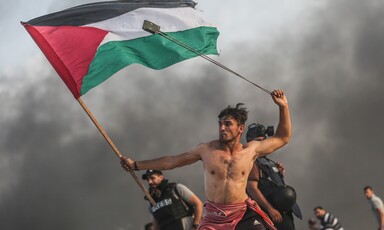 Bare-chested man wields slingshot over his head while holding Palestine flag