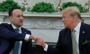 Man wearing tie shakes hands with another man wearing green tie; a white marble fireplace decorated with shamrock and statues can be seen behind them.
