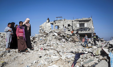 Women and children stand on the remains of a bombed-out building