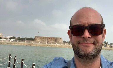 Man wearing sunglasses with body of water and ruins behind him. 