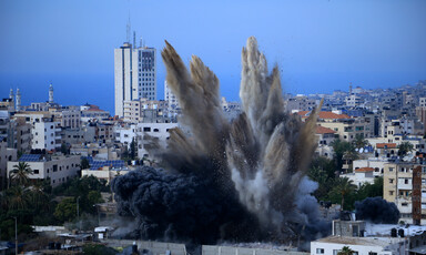 Smoke rises over buildings against Gaza skyline.