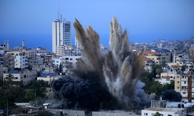 Smoke rises over buildings against Gaza skyline. 