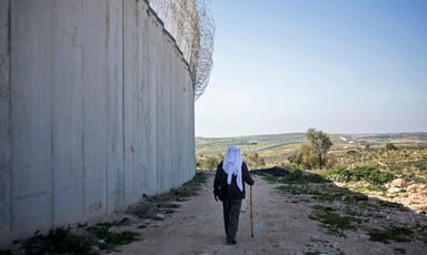 Palestinian man using a cane walks along dirt path next to concrete wall and barbed wire