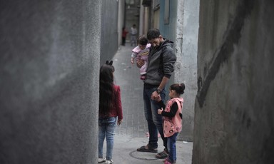 A man carrying a young girl stands flanked by two other children in a narrow grey alley.