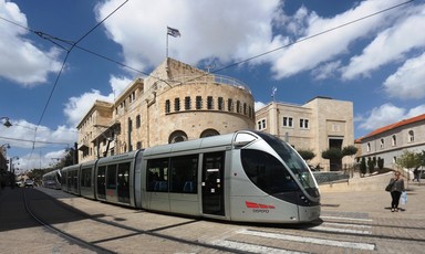 A tram passes through a street with sandstone buildings behind it