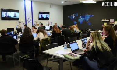 A room full of people working at computers with a large Israeli flag, a world map and monitors on the walls