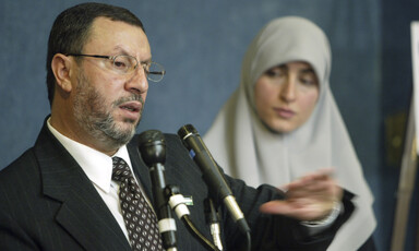 Man speaks before two microphones, woman stands next to him. 