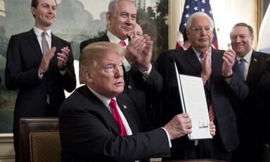 Four standing men applaud another man sat at a desk holding up a piece of paper.