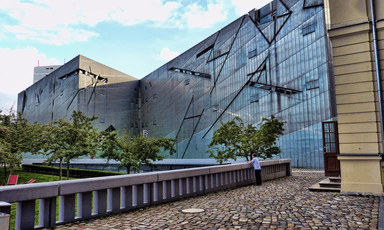 A man is dwarfed by a building that has many angles in its glass facade.