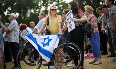 A woman wearing a yellow top holds a bicycle with a blue and white flag draped over it, while talking to another woman holding a placard that reads "Labour: For the Many, Not the Jew." 