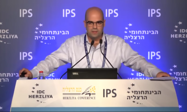 A bald man in a shirt stands at a conference podium speaking into a mic