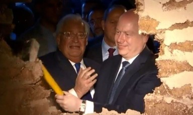 Men in suits, one holding a sledgehammer, look through hole in the wall