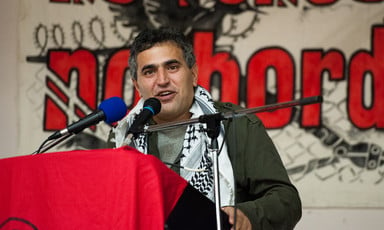 A man wearing a Palestinian scarf speaks at a podium