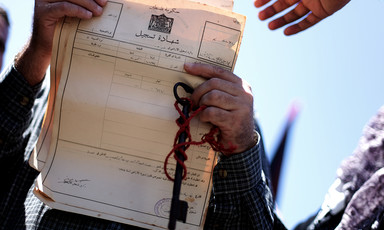 A man's hands with a key and chain wrapped around them holding up a document in Arabic