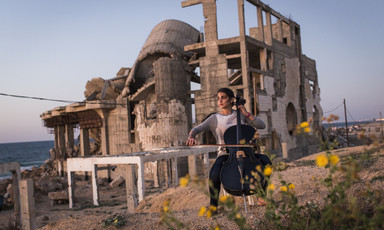 Woman plays cello in front of the rubble of a destroyed building