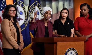 Three women stand around a fourth speaking at podium