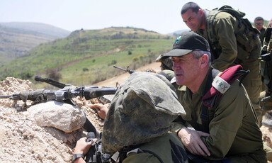 Man in military uniform kneels down next to gun and looks out over hilltops