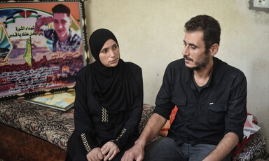 Woman and man sit on a bed; behind them is a large photograph of a boy.