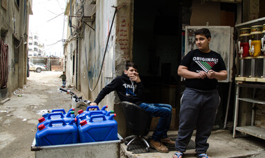 Two boys are next to several large blue plastic water canisters.