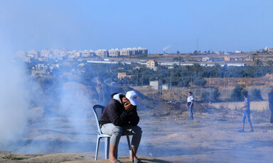 A man sits outside on a plastic chair as smoke billows behind him.