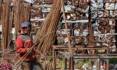 A man carries steel bars with bare hands in a yard filled with scrap metal.