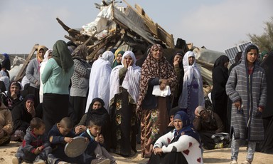 Women and children stand and sit in front of rubble of destroyed structure