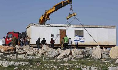 A crane hovers over a prefabricated building, surrounded by large rocks 