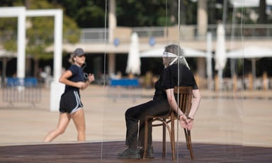 Blindfolded woman cuffed to a chair sits in a glass box 