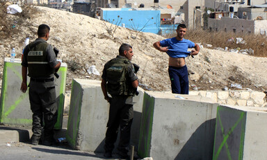 Two soldiers behind concrete blocks look at a young man who has lifted his t-shirt to show his stomach