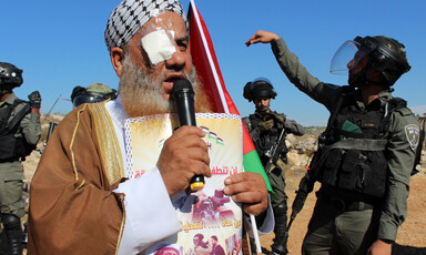 A man with an eyepatch speaks into a microphone while a soldier gesticulates behind him