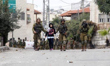 Two armed soldiers walk with a young girl between them while a third points a rifle somewhere in the distance