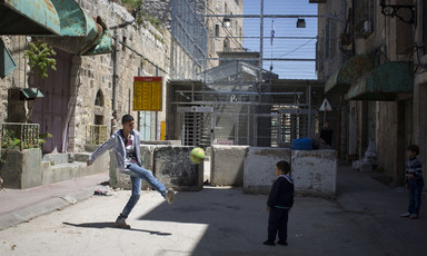 Two boys play with soccer ball in front of concrete blocks and metal barricade