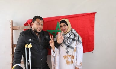 A couple flash the victory sign in front of a Moroccan flag.