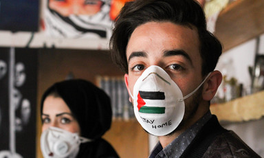 A man and a woman wear painted face masks one bearing the Palestinian flag and  words stay at home