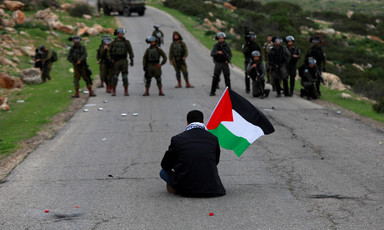 A man holding a Palestine flag sits on a road facing a line of soldiers