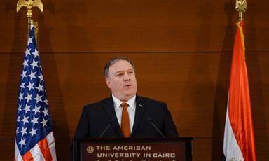 Man in suit stands before podium between two flags 