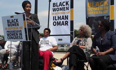 Woman speaks at podium with women and banners behind her