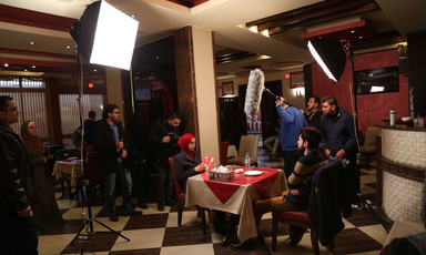 Film crew members stand around male and female actors sitting on opposite sides of a restaurant table