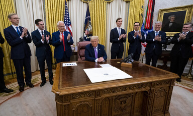 Man sits behind desk surrounded by men